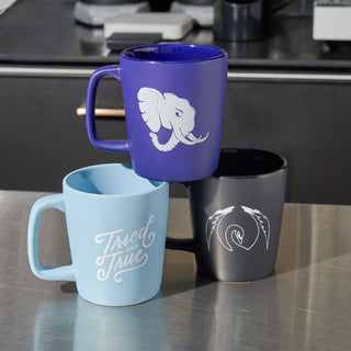 Purple coffee mug with elephant graphic stacked on top of blue tried and true mug and black coffee mug with a black swan graphic.