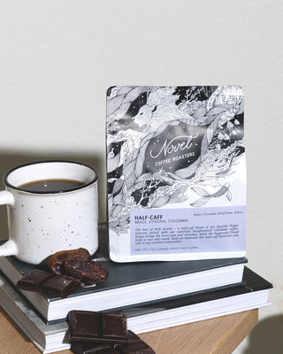 Half Caffeinated bag of coffee on top of books, next to a cup of brewed coffee, bakers chocolate, and dates.